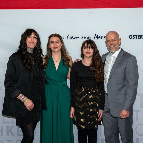 Vier Menschen stehen eng beieinander auf einem roten und weißen Teppich und lächeln für ein Foto. Eine Frau links trägt ein schwarzes Kleid und eine Jacke. Eine Frau in der Mitte trägt ein grünes Kleid. Eine Frau rechts trägt ein schwarzes und goldenes Kleid. Ein Mann rechts trägt einen grauen Anzug.