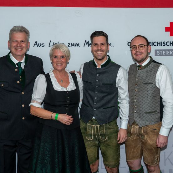 Vier Personen, zwei Männer und zwei Frauen, gekleidet in traditioneller Kleidung, posieren für ein Foto vor einem weißen Hintergrund mit einem roten Kreuz.