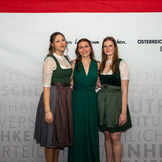 Drei Frauen stehen gemeinsam vor einem weißen Hintergrund mit Text. Die mittlere Frau trägt ein grünes Kleid, während die anderen grüne Kleider mit weißer Spitze tragen. Sie scheinen für ein Foto zu lächeln.