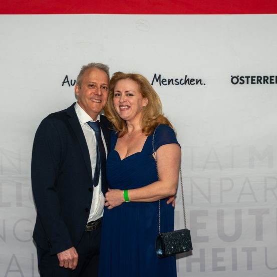 Ein lächelndes Paar steht eng beieinander und posiert für ein Foto. Der Mann in Anzug und Krawatte, die Frau in einem blauen Kleid mit einem grünen Armband, vor einer Wand mit Text.
