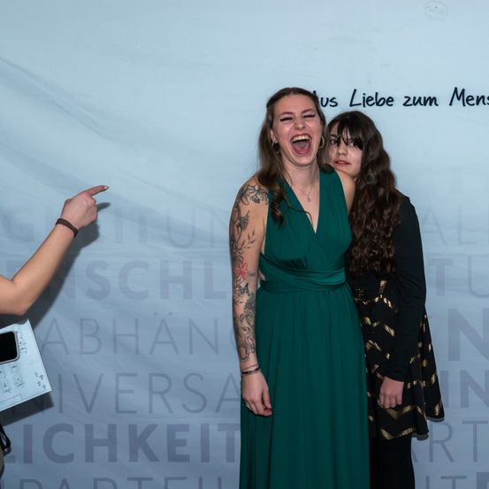 Zwei Frauen stehen zusammen, eine lacht. Sie sind bei einer Veranstaltung, mit einem weißen Hintergrund dahinter. Die Frau auf der linken Seite trägt ein grünes Kleid und hat Tattoos. Eine andere Frau steht neben ihr, trägt ein schwarzes und goldenes Kleid.
