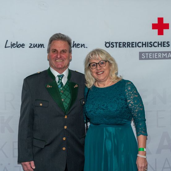 Ein älteres Paar steht lächelnd für ein Foto vor einer weißen Wand mit dem Text 'Liebe zum hen.' Der Mann trägt eine formelle grüne und goldene Jacke mit Krawatte, und die Frau trägt ein blaues Kleid und eine Brille. Hinter ihnen befindet sich ein rotes Kreuz-Logo.