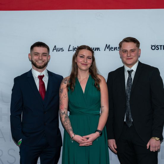 Drei Menschen in formeller Kleidung stehen gemeinsam, lächeln für ein Foto. Der Mann links trägt eine rote Krawatte, die Frau in der Mitte ein grünes Kleid und der Mann rechts einen schwarzen Anzug. Sie stehen vor einer Wand mit deutschem Text.