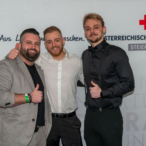 Drei Männer stehen zusammen vor einem weißen Hintergrund und lächeln für ein Foto. Der Mann auf der linken Seite gibt einen Daumen hoch. Der Mann in der Mitte trägt ein weißes Hemd und schwarze Hose. Der Mann auf der rechten Seite trägt ein schwarzes Hemd, eine Hose mit Hosenträgern und eine schwarze Hose.