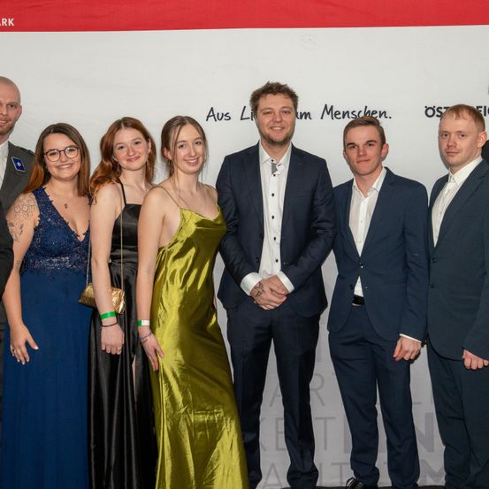 Eine Gruppe von Menschen in formeller Kleidung, darunter drei Frauen und vier Männer, posiert für ein Foto vor einem Hintergrund mit Text auf Deutsch.