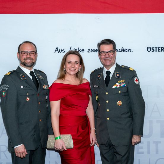 Drei Personen stehen vor einem weißen Hintergrund, zwei Männer in Militäruniformen und eine Frau in einem roten Kleid. Sie alle lächeln und scheinen für ein Foto zu posieren.