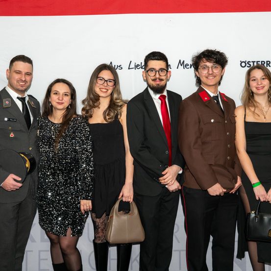 Eine Gruppe von sechs Menschen in formeller Kleidung posiert für ein Foto vor einem weißen Hintergrund mit Text. Zwei Männer in Anzügen und Krawatten und eine Frau in Uniform sind anwesend.