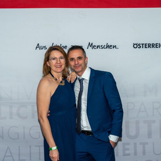 Ein Mann und eine Frau lächeln für ein Foto. Der Mann trägt einen blauen Anzug und eine Krawatte. Die Frau trägt ein blaues Kleid. Sie stehen vor einer Wand mit Schrift.
