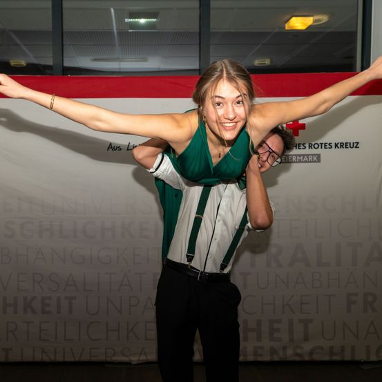 Eine lächelnde Frau in einem grünen Kleid wird von einem Mann in einer Brille hochgehoben, mit einem großen Hintergrund vor ihnen, auf dem das Rote-Kreuz-Logo und Text zu sehen sind.