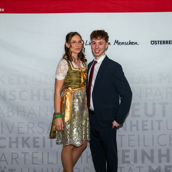 Ein junges Paar steht lächelnd vor einem Banner mit deutschem Text. Die Frau trägt ein traditionelles Kleid, der Mann ist formell gekleidet.