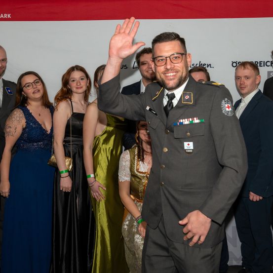 Ein Mann in Uniform winkt zur Kamera, während mehrere Personen in formeller Kleidung bei einer Veranstaltung hinter ihm stehen.