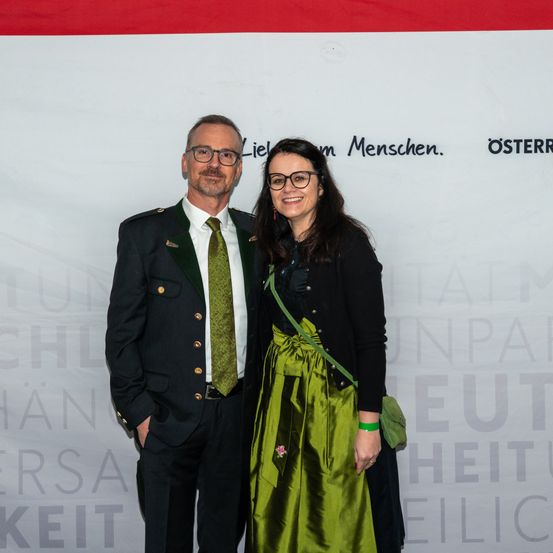 Ein erwachsener Mann und eine Frau stehen vor einem weißen Hintergrund mit Text, lächeln und schauen in die Kamera. Der Mann trägt eine schwarze Uniform mit einer grünen Krawatte, und die Frau trägt ein grünes Kleid.