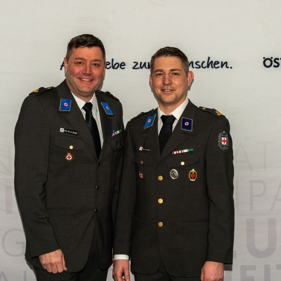 Zwei Männer in Militäruniformen stehen nebeneinander vor einem weißen Hintergrund mit deutschem Text. Sie lächeln und posieren für ein Foto.