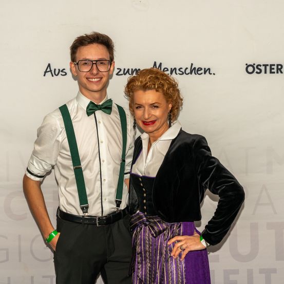 Ein Mann und eine Frau stehen gemeinsam für ein Foto. Der Mann trägt eine Brille und Hosenträger. Die Frau ist in einem schwarzen und lilafarbenen Outfit gekleidet. Sie lächeln beide in die Kamera. Hinter ihnen befindet sich eine Wand mit Text in deutscher Sprache.