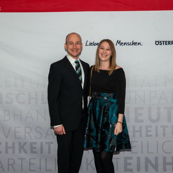 Ein Mann und eine Frau stehen eng beieinander vor einem Banner mit den Worten Liebe Menschen. Osterr. Sie lächeln beide und scheinen für ein Foto zu posieren.