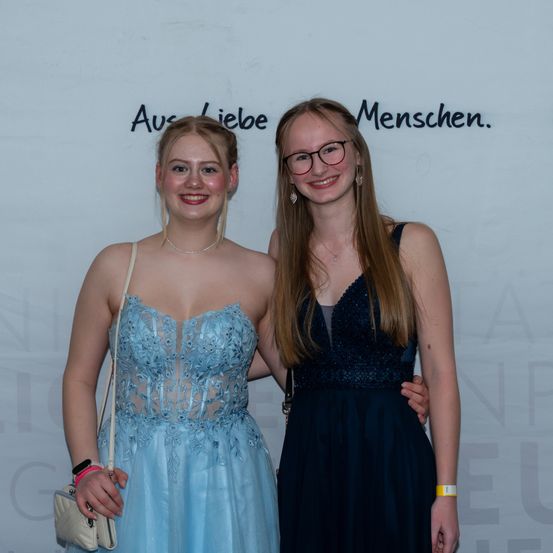 Zwei junge Frauen stehen eng nebeneinander vor einer Wand mit dem Text 'Aus Liebe Menschen.' Sie lächeln beide für ein Foto.