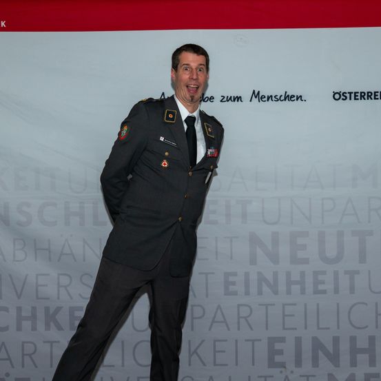 Ein Mann in Uniform steht mit einem freudigen Ausdruck. Hinter ihm befindet sich ein großes weißes Banner mit verschiedenen Texten und Logos, darunter 'Osterreich' und 'be zum Menschen'.