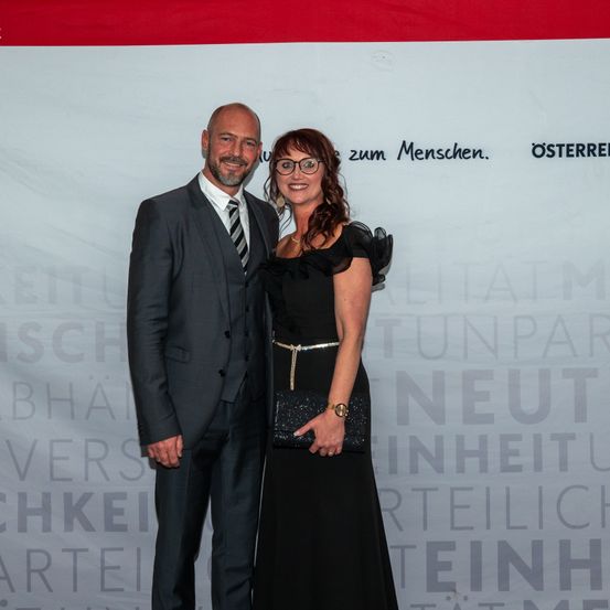 Ein Mann und eine Frau stehen lächelnd nebeneinander in formeller Kleidung. Der Mann trägt einen grauen Anzug mit gestreifter Krawatte, und die Frau trägt ein schwarzes Kleid mit einem Gürtel. Sie stehen vor einem Banner mit verschwommenem Text.