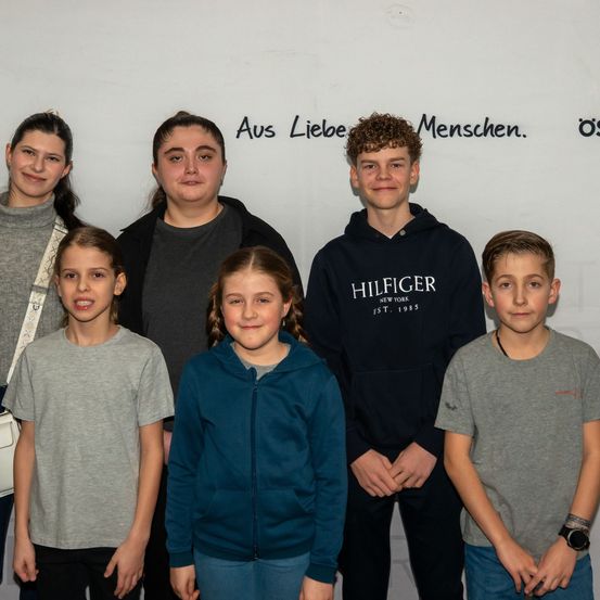 Eine Gruppe von Kindern und jungen Erwachsenen posiert für ein Foto vor einer weißen Wand mit den Worten 'Aus Liebe Menschen'.