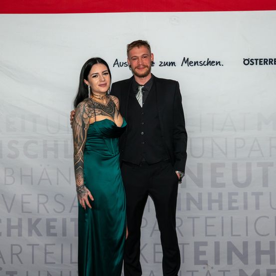 Ein Mann und eine Frau stehen lächelnd bei einer Veranstaltung. Die Frau hat Tattoos an den Armen und trägt ein grünes Kleid. Der Mann ist in einem schwarzen Anzug und einer Krawatte. Sie stehen vor einem Banner mit weißem Text.