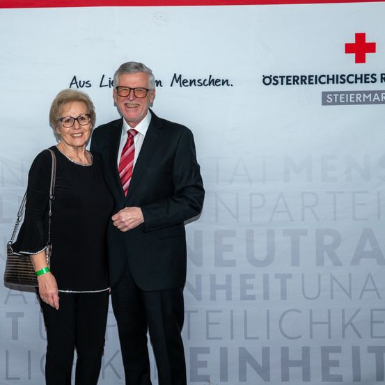 Ein älteres Paar steht eng beieinander vor einer Wand mit dem Text 'Aus Ligen Menschen.' Der Mann trägt einen Anzug und eine Krawatte, und die Frau hat eine Handtasche. Hinter ihnen befindet sich ein großes rotes Kreuz.