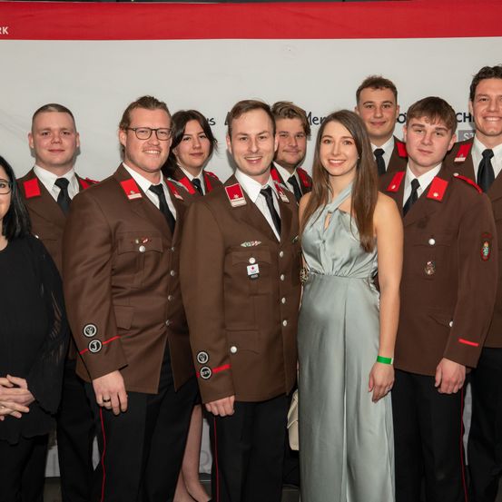 Eine Gruppe von Menschen in braunen Uniformen mit roten Abzeichen steht zusammen, wahrscheinlich bei einer formellen Veranstaltung. Eine Frau in einem grauen Kleid steht in der Mitte.