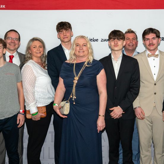 Eine Familie steht vor einem weißen Banner mit rotem Text. Eine Frau in einem blauen Kleid hält eine Handtasche. Zwei Männer tragen Anzüge, einer mit einer Fliege. Ein Mann trägt eine rote Krawatte. Zwei Frauen tragen Halsketten. Zwei Männer tragen eine Brille.