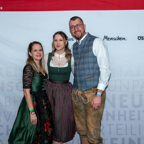 Drei Personen stehen eng beieinander vor einem weißen Banner mit Text. Der Mann trägt eine Weste und eine Brille, und die Frauen tragen traditionelle grüne Kleider.