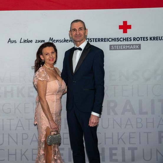 Ein Mann und eine Frau in formeller Kleidung stehen gemeinsam auf einem roten Teppich. Sie lächeln für ein Foto vor einer Kulisse mit dem Roten Kreuz-Symbol und Text.