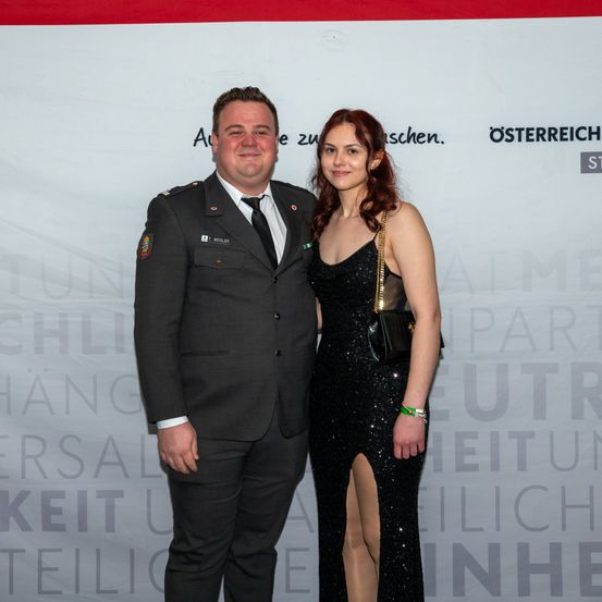 Ein Mann in Uniform und eine Frau in einem schwarzen Kleid stehen nebeneinander und lächeln. Sie stehen vor einem weißen Hintergrund mit schwarzem Text.