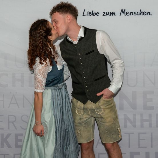 Ein Mann und eine Frau küssen sich auf dem roten Teppich. Die Frau trägt ein traditionelles Kleid und der Mann trägt eine Weste und Shorts. Hinter ihnen ist ein weißer Hintergrund mit deutschem Text.