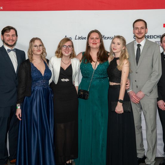 Eine Gruppe von sechs Personen posiert für ein Foto bei einer formellen Veranstaltung. Sie stehen vor einem Banner.