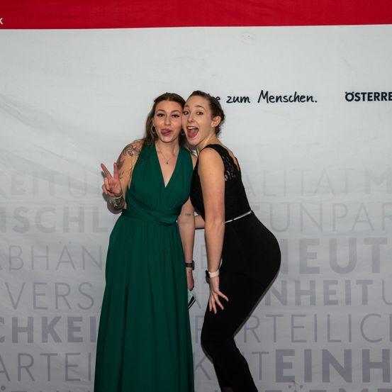 Zwei Frauen posieren für ein Foto, eine in einem grünen Kleid und die andere in einem schwarzen Kleid, lächeln und machen lustige Gesichter. Hinter ihnen ist ein weißer Hintergrund mit Text.