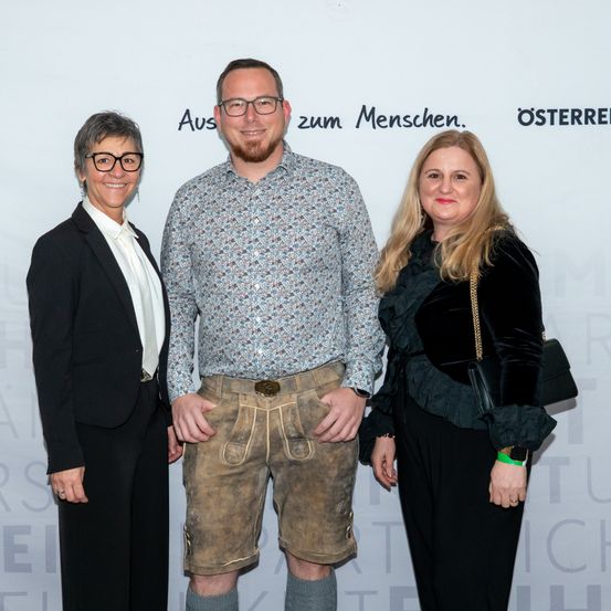 Drei Personen stehen vor einer Wand mit einem Slogan. Ein Mann in traditioneller bayerischer Kleidung wird von zwei Frauen flankiert. Sie alle lächeln in die Kamera.