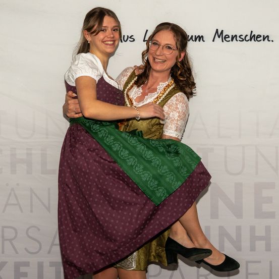 Zwei Frauen in traditioneller bayerischer Kleidung, eine in einem Dirndl und die andere in einer Spitzenbluse, umarmen sich fröhlich und lächeln vor einer weißen Wand.