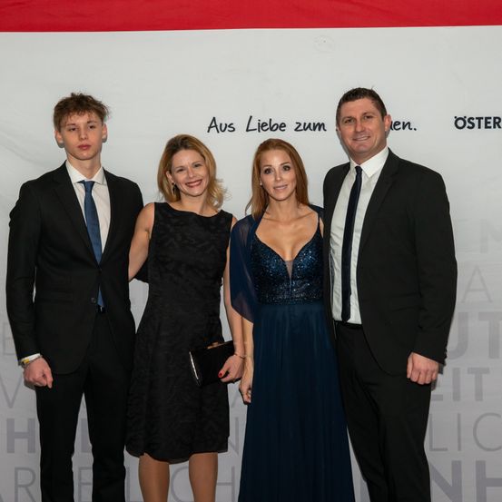Vier Personen stehen zusammen in formeller Kleidung und lächeln für ein Foto. Die beiden Frauen tragen Kleider, eines blau und eines schwarz. Die Männer tragen Anzüge. Hinter ihnen befindet sich ein weißer Banner mit deutschem Text.