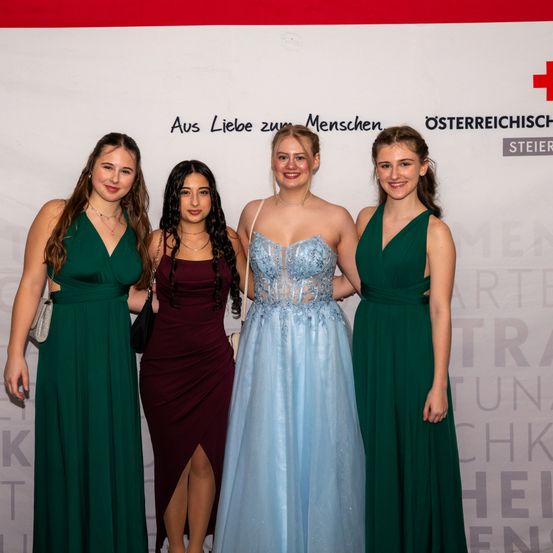 Vier Frauen stehen gemeinsam auf einem roten Teppich. Eine Frau trägt ein blaues Kleid, während die anderen grüne Kleider tragen. Sie alle lächeln und schauen in die Kamera. Hinter ihnen ist eine weiße Wand mit Text und einem roten Kreuz-Logo.