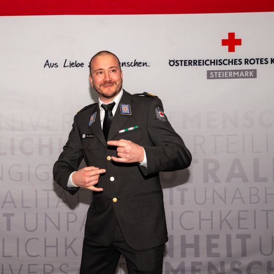 Ein Mann in Uniform mit einem roten Kreuz-Symbol steht vor einem Banner. Das Banner hat Text auf Deutsch, einschließlich 'Aus Liebe Menschen'. Er lächelt und gestikuliert mit den Händen.