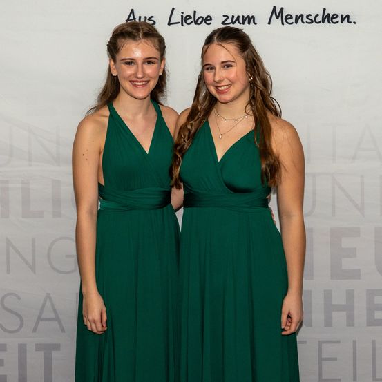 Zwei Frauen in grünen Kleidern stehen nebeneinander und lächeln für die Kamera. Sie sind bei einer Veranstaltung mit einem weißen Hintergrund.
