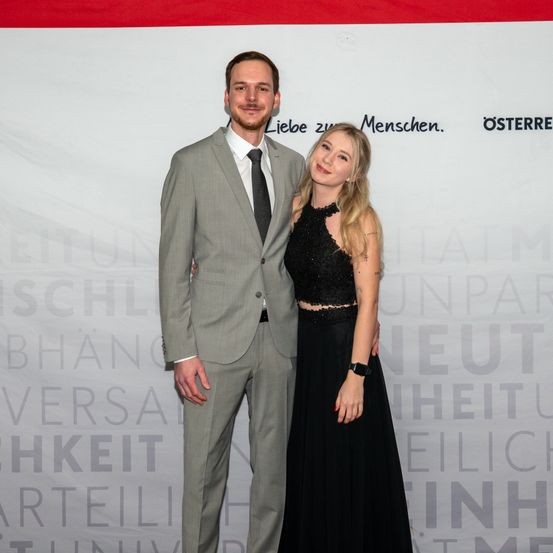 Ein Mann und eine Frau stehen nah beieinander auf einem roten Teppich und lächeln. Der Mann trägt einen grauen Anzug und die Frau ein schwarzes Kleid. Hinter ihnen ist ein weißer Hintergrund mit verschwommenem Text.
