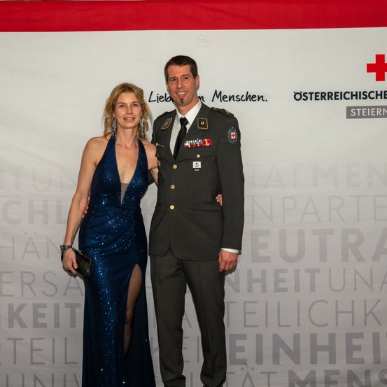 Ein Mann in Uniform steht neben einer Frau in einem blauen Paillettenkleid und lächelt für ein Foto vor einem Hintergrund mit Text.