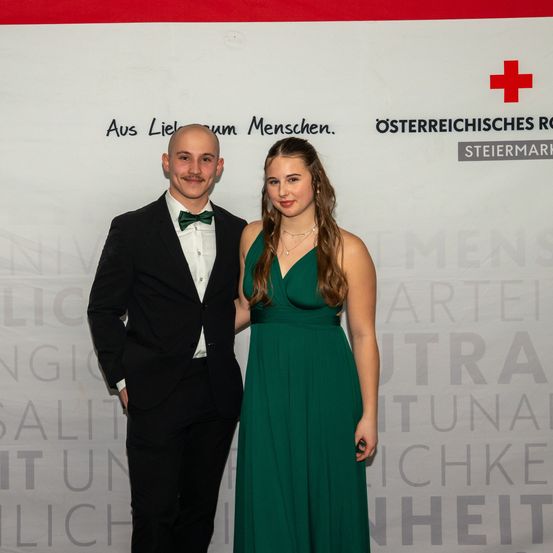 Ein Mann und eine Frau stehen lächelnd vor einer weißen Wand mit Text und posieren für ein Foto. Der Mann trägt einen schwarzen Anzug und eine grüne Fliege, während die Frau ein grünes Kleid trägt. Hinter ihnen ist ein rotes Kreuz sichtbar.