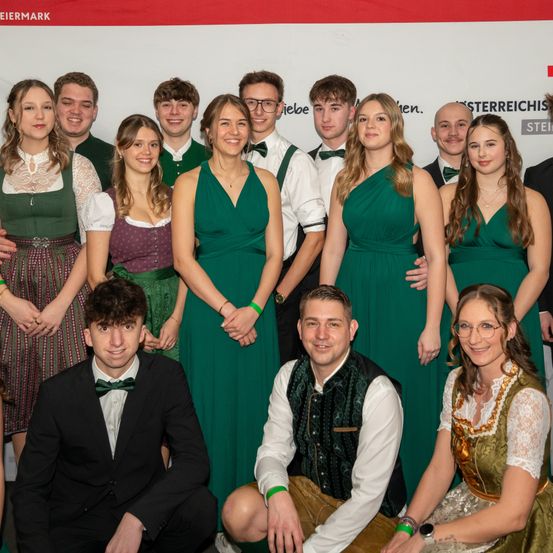 Eine Gruppe von Menschen in traditioneller deutscher Kleidung, die für ein Foto posiert. Im Hintergrund ist ein Banner mit dem Wort 'Sterreich' zu sehen.