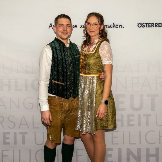 Ein Mann und eine Frau in traditioneller bayerischer Kleidung stehen eng beieinander und lächeln für die Kamera, mit einer Wand im Hintergrund.