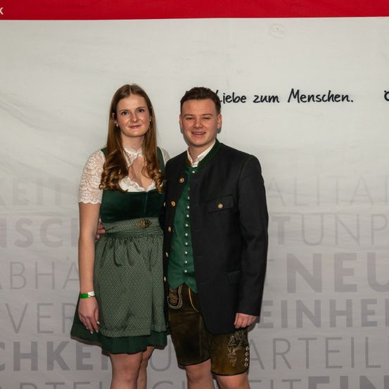 Ein junges Paar steht nah beieinander und lächelt in traditioneller deutscher Kleidung. Sie stehen vor einem Banner mit Text in Deutsch.