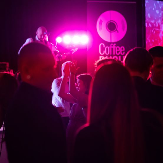 Eine Band spielt auf der Bühne in einem schwach beleuchteten Veranstaltungsort. Eine Frau im Publikum tanzt. Das Wort Kaffee ist auf einem Banner sichtbar.