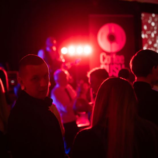 Eine Gruppe von Menschen ist auf einer Party mit roten Lichtern und einem großen Schild, das Coffee Rush liest. Einige Leute stehen, während andere sitzen.