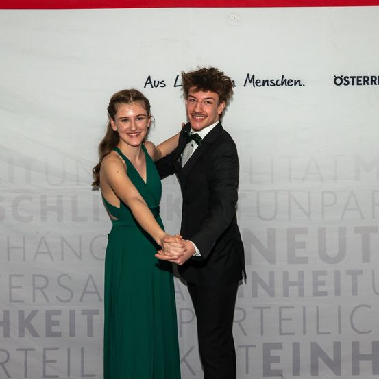 Ein junges Paar in formeller Kleidung tanzt vor einer Wand mit Worten wie 'Aus Menschen gemacht' und 'Österreich'. Die Frau trägt ein grünes Kleid, und der Mann ein schwarzes Anzug.