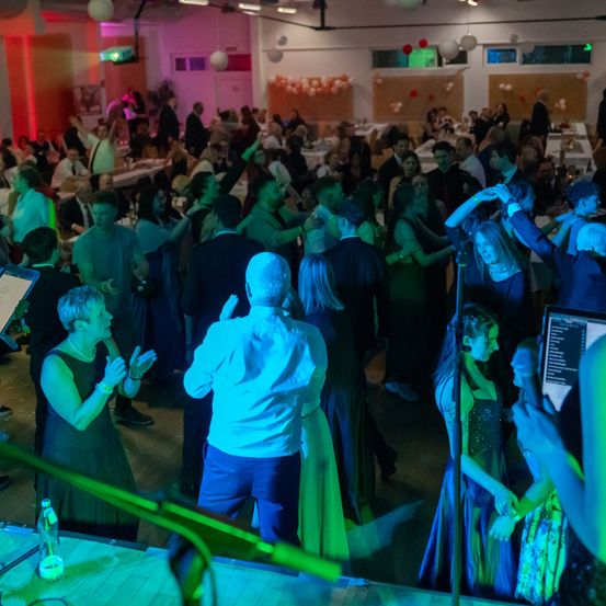 Eine lebhafte Tanzfläche mit Menschen in formeller Kleidung, die unter Disco-Lichtern tanzen. Einige stehen auf der Bühne, während andere im Hintergrund tanzen, mit Dekorationen wie Luftballons und Lichtern.