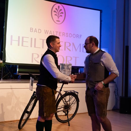 Zwei Männer in traditioneller bayerischer Kleidung schütteln sich die Hände vor einer Bühne. Ein Fahrrad steht neben ihnen. Eine große Leinwand zeigt 'Bad Waltersdorf Heiltherme Resort' an.